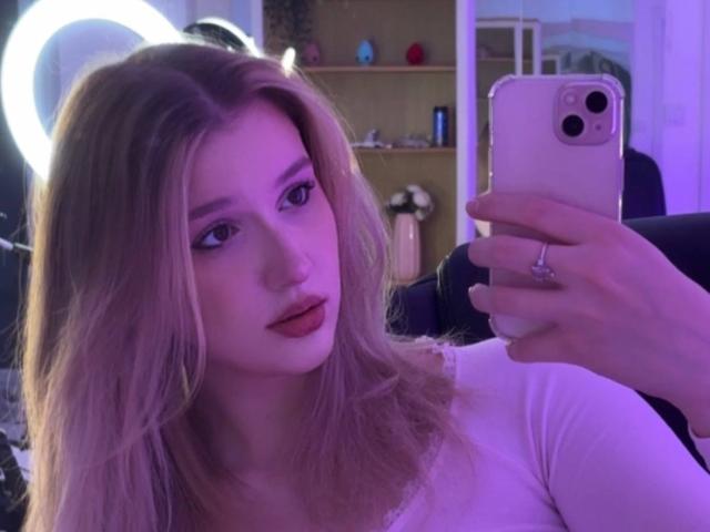 Synryx mit blonden Haaren, in einem rosa-lila Licht, hält ein Telefon, macht ein Selfie, trägt ein weißes Oberteil, hat einen Ring am Finger, schaut mit geschürzten Lippen in die Kamera..