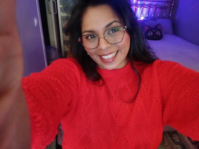 Chili with dark hair, glasses, and red knitted sweater, smiling at the camera, showing her teeth, holding her phone in the foreground, purple room in the background.