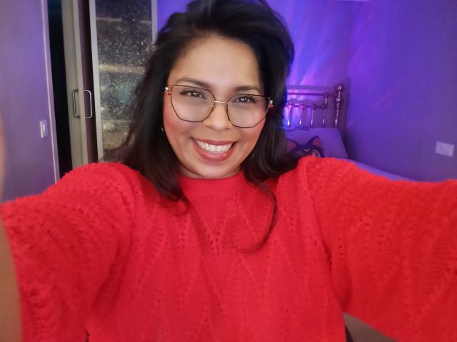 Chili with dark hair, glasses, and red sweater, smiling at the camera. She has medium tits, pink lipstick, and is taking a selfie in a purple-lit room.