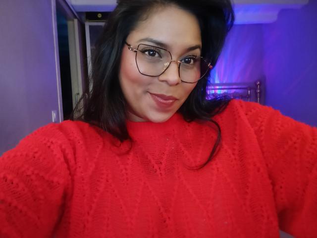 Chili, with dark hair and glasses, wearing a red, textured sweater, smiling at the camera. Her tits are small, and she has a cute, confident look.