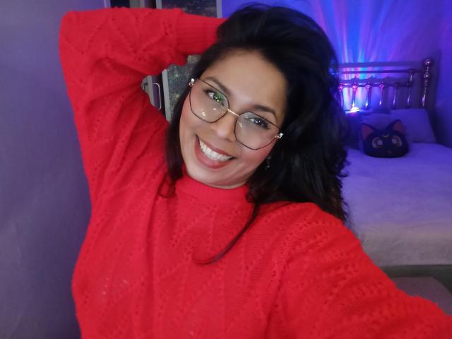 Chili with big tits, wearing a red knit sweater, smiling at the camera, arm raised, glasses on, black wavy hair, blue eyes, and purple room lighting in the background.
