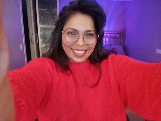 Chili with medium brown hair, glasses, and a red textured sweater, smiling at the camera, holding up her right arm. Her tits are partially visible under the sweater.