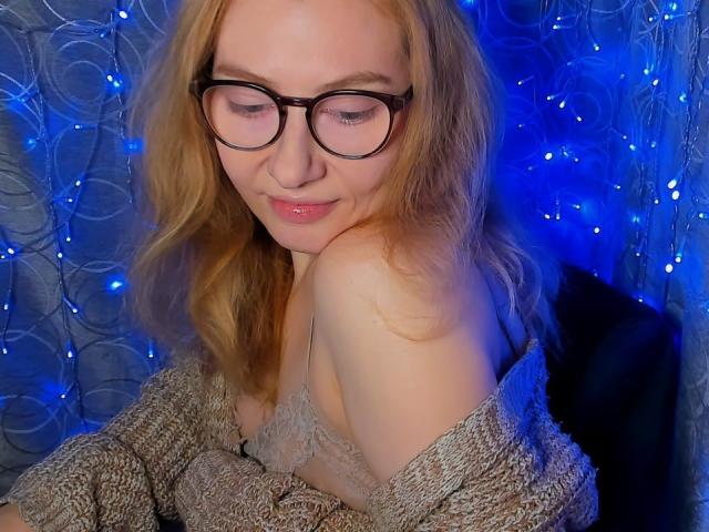 PaulinaSunny with glasses, red hair, and a cute smile, wearing a lace bra and a knitted sweater, showing her bare shoulder and a subtle hint of her tits.