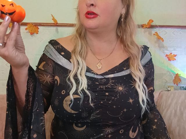 KelliRedhaer holding a pumpkin in her right hand, wearing a black, celestial-patterned dress with grey sleeves, and red lipstick, with blonde hair and a gold necklace.