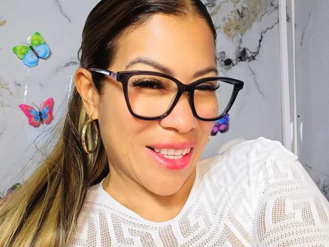 AlixLow, avec ses lunettes noires, son rouge à lèvres rose, sa chemise blanche en dentelle et ses boucles d'oreilles cerceaux, sourit à l'appareil photo. On peut voir des décorations murales en forme de papillons. Ses cheveux bruns sont attachés en arrière..