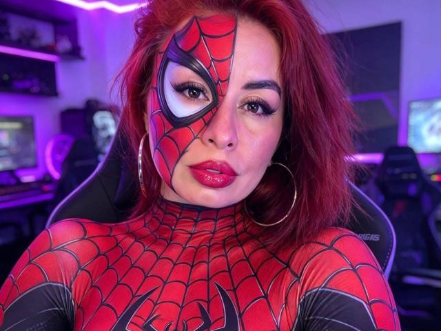 IvvannaParis with red hair, wearing a Spider-Man mask and red, web-patterned bodysuit, hoop earrings, and glossy red lips, looking directly at the camera, in a dimly lit room.