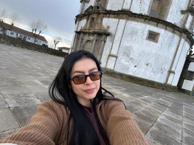 IsabellaJames with long black hair, brown sweater, and black sunglasses, smiling at the camera, standing in front of a worn, white, historic building with a stone courtyard.