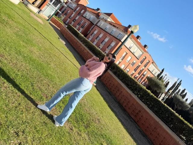 IsabellaJames stands on grass, leaning against a brick wall, wearing a pink sweater, blue jeans, and white sneakers, with a blue sky and red-brick building in the background.