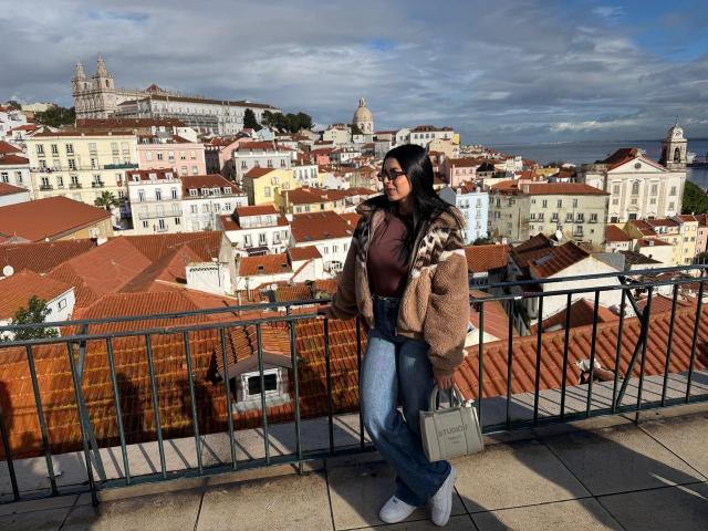 IsabellaJames with brown fur coat, black hair, sunglasses, blue jeans, white heels, and gray purse, standing on balcony, looking left, cityscape with red roofs and historic buildings.