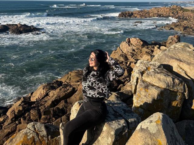 IsabellaJames with dark hair, wearing glasses, black-and-white patterned shirt, and black pants, sitting on rocky shoreline, looking at camera, ocean waves in background, sunny day.