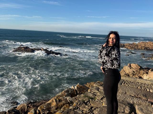 IsabellaJames stands on rocky shoreline, wearing a black and white patterned top with black pants, her dark curly hair blowing in the wind, ocean waves in the background.