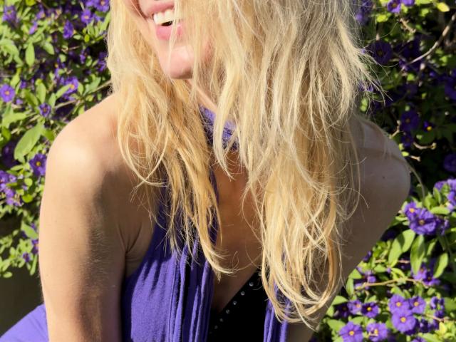 ShaiyaShy-hot with wavy blonde hair, purple dress, and smiling mouth, showing bare shoulders and a hint of cleavage, surrounded by vibrant purple flowers.