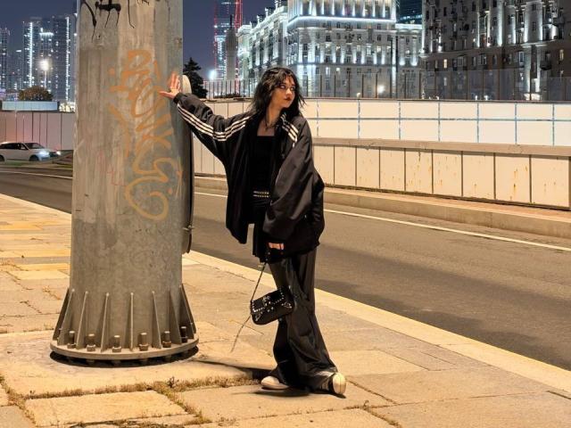 LakeshiaPronto est appuyée contre un poteau recouvert de graffitis, vêtue d'une veste noire, d'un pantalon ample et de baskets blanches, avec une sangle d'appareil photo pendue à sa main. Paysage urbain nocturne..