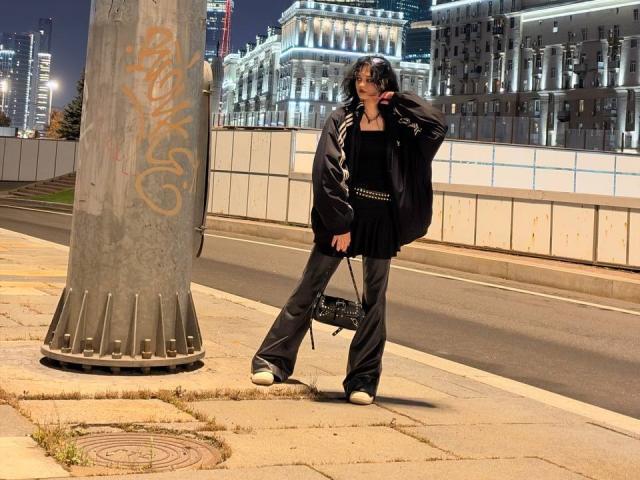 LakeshiaPronto se tient sur un trottoir la nuit, vêtue d'une veste noire, d'une robe noire et d'un pantalon évasé, tenant un sac noir, avec des graffitis sur un poteau à côté d'elle..