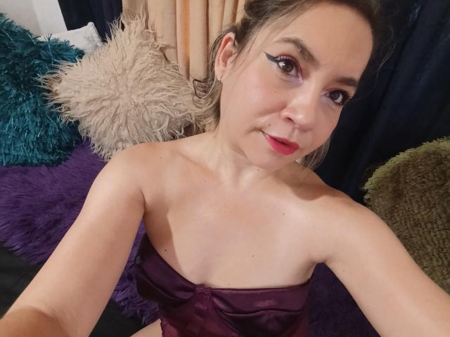 MsJustineBeaux, con piccoli seni, indossa una top viola in raso senza spalline, ha la pelle chiara, i capelli castani e il rossetto rosso, e guarda la fotocamera con un leggero sorriso..
