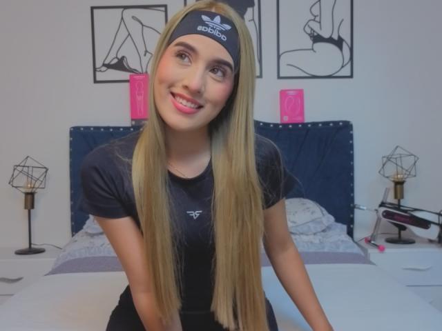 EmmaWat with long blonde hair, black Adidas headband, black t-shirt, smiling, sitting on bed, pink nose ring, cute, bed with blue headboard, white walls, geometric lamps.