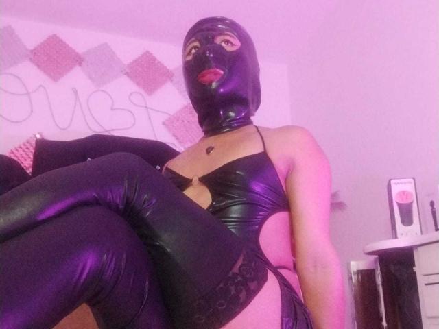 JatziBehot69 wearing a black latex mask, shiny black leather bra, and matching thigh-highs, sitting with legs crossed, showing a sexy cleavage and black lace garter on her right thigh.