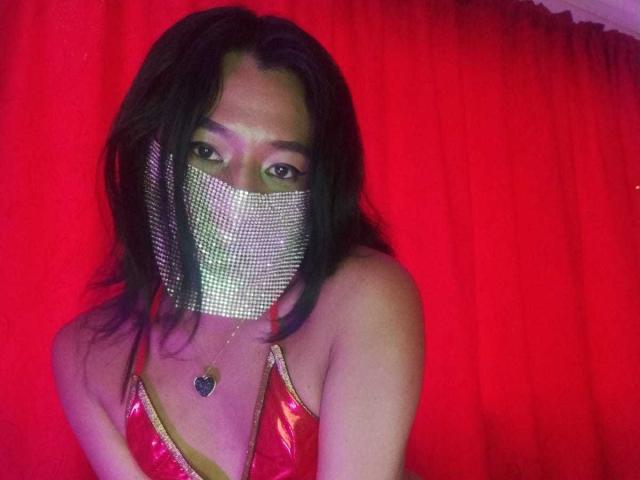 JatziBehot69 with long black hair, wearing a shiny red dress and a glittery face mask, and a heart-shaped necklace, looking at the camera with sultry eyes.