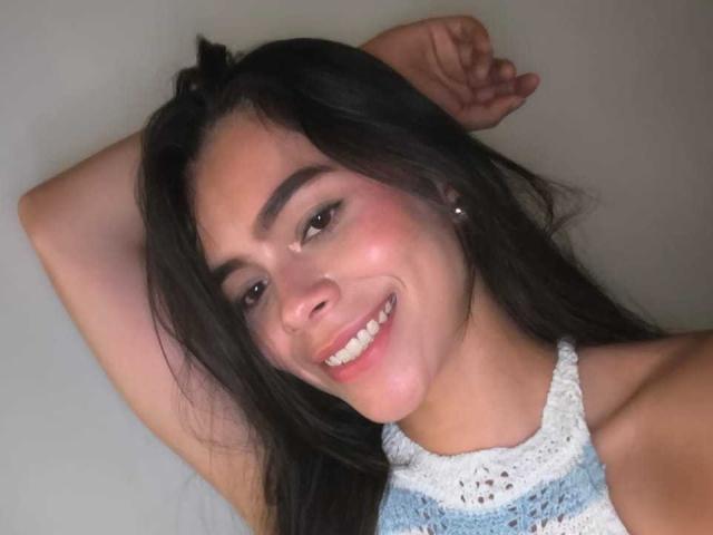 AlanaBlake has long dark hair, brown eyes, and a cute smile. She's wearing a white and blue crocheted top, with a small nose piercing and a relaxed, sexy vibe.