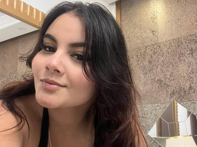 ReginaMills has dark, wavy hair, a small nose, and full lips with a slight smile. She's wearing a black top, showing her tits in the photo.