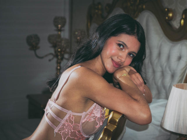 AmilyPreston with dark wavy hair, pink lace bra, and a smile, resting her chin on a gold chair, showing her tits and a gold bracelet. Soft, dim bedroom light.