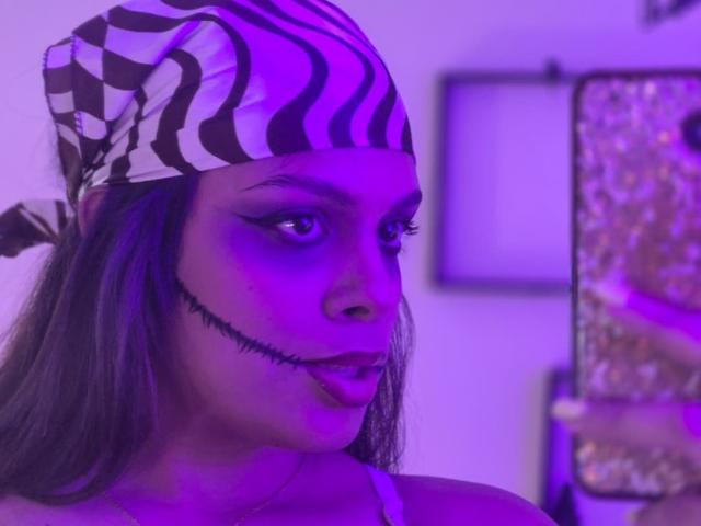 TeylorJenkins with zebra-pattern headscarf, purple-blue lighting, dark eyeliner, black stitched mouth, long brown hair, holding glittery phone, tongue slightly out, looking at camera.