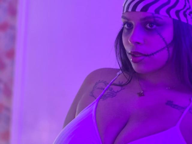 TeylorJenkins with zebra-pattern headband, purple-lit face, black tattoo under right eye, white tank top showing big tits, necklace with small heart, looking to the side.
