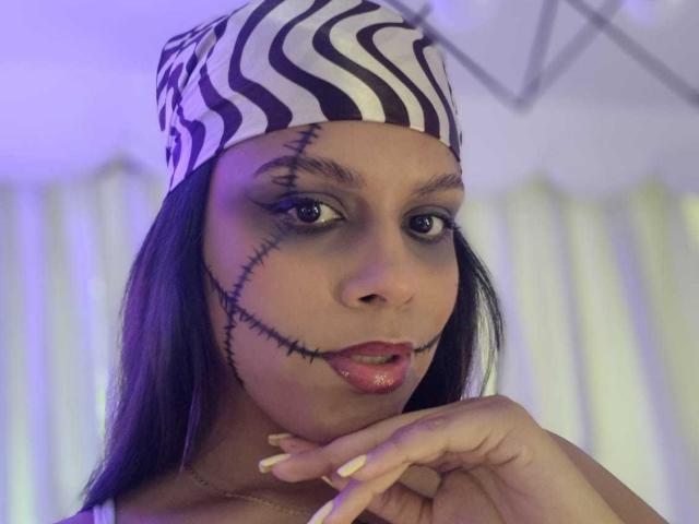 TeylorJenkins with dark skin, black hair, wearing a black-and-white striped hat with a stitched face, red lipstick, and a gold necklace, touching her chin.