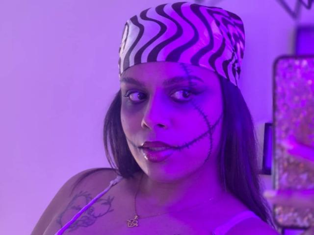 TeylorJenkins with a zebra-patterned cap, black eyeliner, and stitches on her face, purple-lit, showing tattoos on her shoulder, and a necklace with a small pendant.