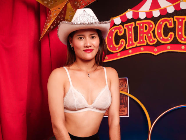 AmberGarcias with medium tits, wearing a white bra, cowboy hat, black skirt, and red lipstick, standing in front of a circus-themed backdrop.