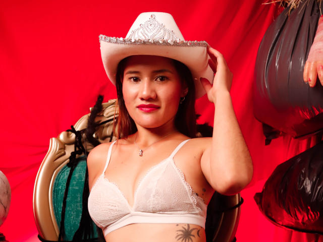 AmberGarcias with brown hair, white lace bra, and cowboy hat, holding hat, showing tits, tattoo on side, red background, smiling, green and gold chair, hanging black bags.