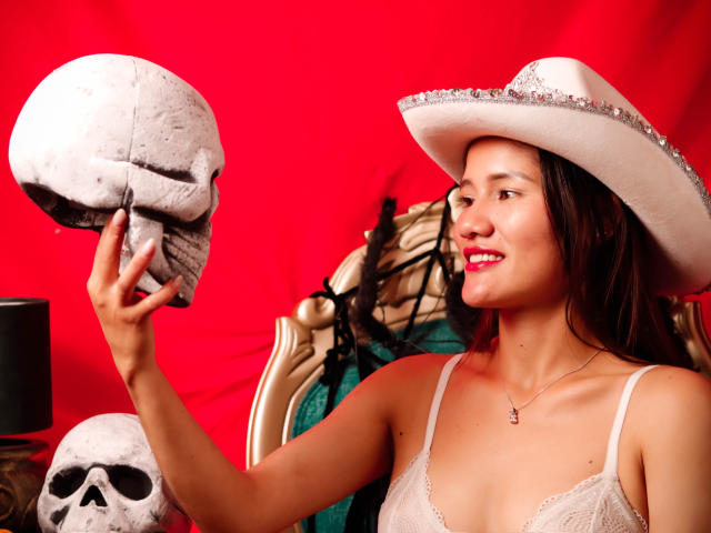 AmberGarcias holding a skull with her right hand, wearing a white lace top and a sparkly white hat, smiling, with red background and another skull on the left.