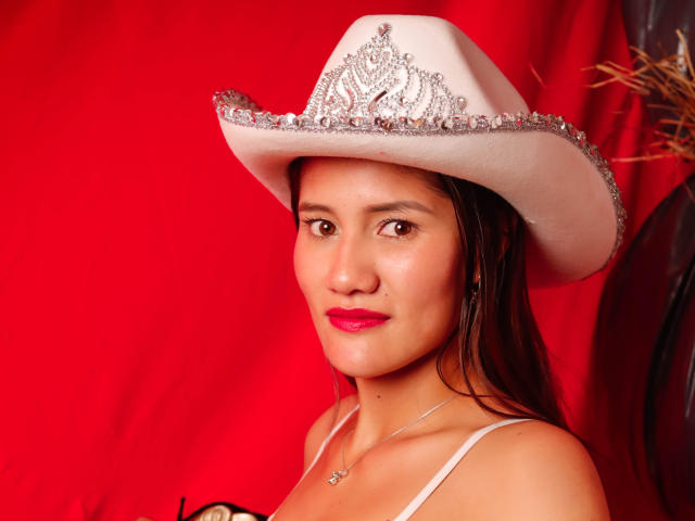 AmberGarcias with brown eyes, red lipstick, wearing a white cowboy hat with silver embellishments, and a white strap top, showing her tits and a cute smile.