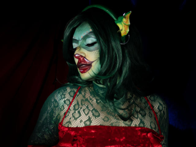 KatParadise with green headband, dark wavy hair, white makeup, red lipstick, green eyes, red velvet top, lace sleeves, mouth open, dark background, red and black fabric.