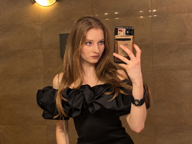 ArdelleCooksey with long brown hair, black off-shoulder dress, holding phone in mirror selfie, brown tiled wall, looking serious, tattoo on right arm, wristwatch on left.