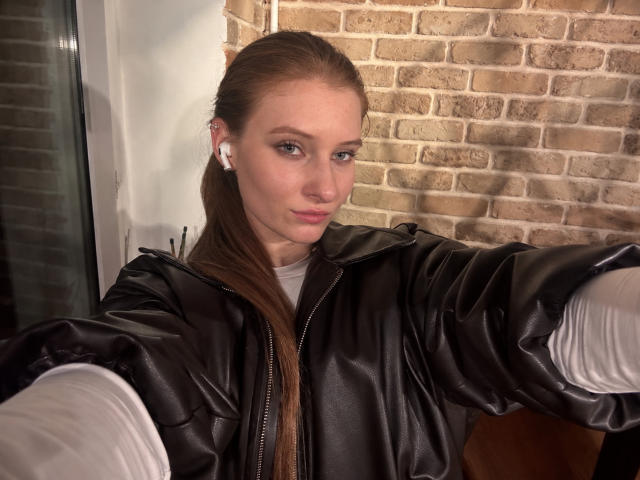 ArdelleCooksey with long brown hair, wearing a black leather jacket, white earbuds, and white shirt, taking a selfie, showing off her pouty face and slight smirk.