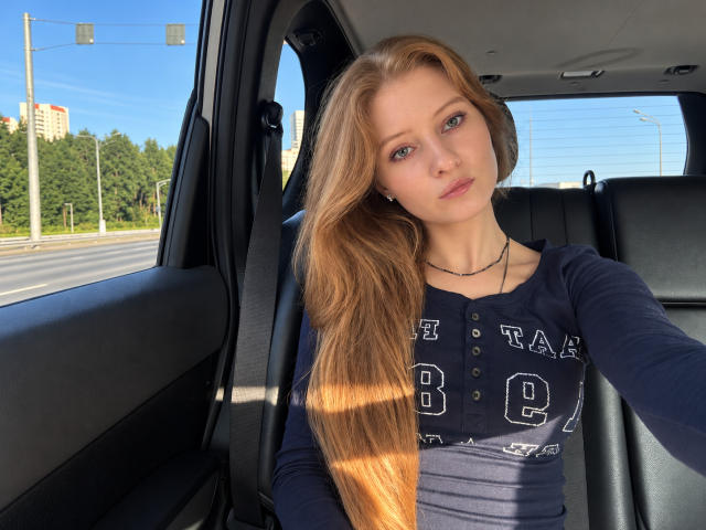 ArdelleCooksey with long, straight, red-brown hair, wearing a navy blue, button-up, long-sleeve shirt, sitting in a car, taking a selfie, showing her tits slightly, looking at camera.