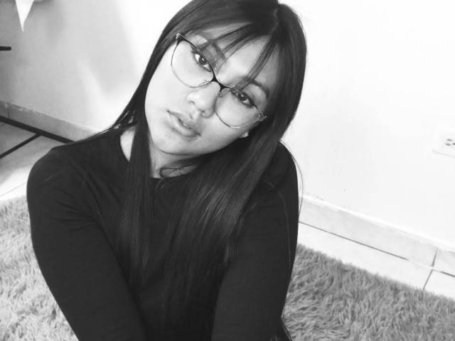 Catalehia69 with long dark hair, glasses, and pouty lips, wearing a black long-sleeve shirt, sitting on a fluffy rug, looking up at the camera. Black-and-white selfie.