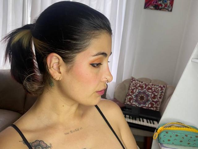 NyxVale with dark hair in a ponytail, nose ring, and pink lipstick, looking down, showing her small tits and a tattoo on her chest, in a brightly lit room.