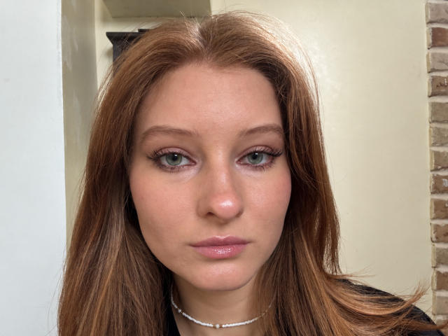 ArdelleCooksey has light brown hair, green eyes, and a neutral expression with pink lips. She's wearing a black top and a silver necklace. Her skin is fair.