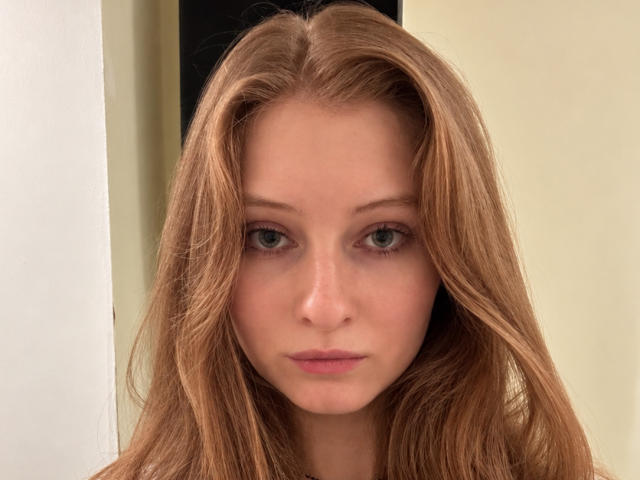 ArdelleCooksey has long, straight, blonde hair and green eyes, with a neutral expression on her slightly pinkish lips, looking straight into the camera. Her face is centered.