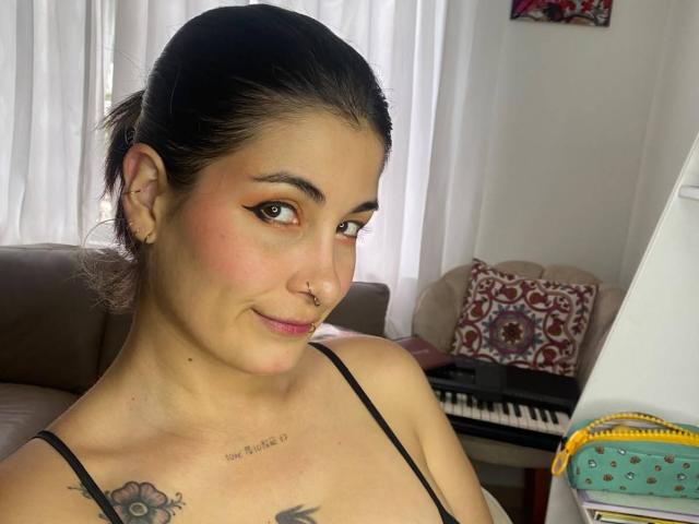 NyxVale has dark hair in a ponytail, brown eyes, and a slight smile. She's wearing a black spaghetti strap top, showing a tattoo on her chest and nose ring.