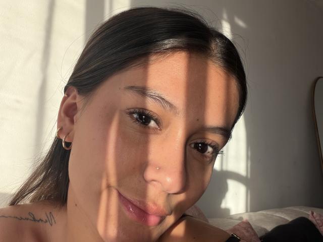 Thalia Ares tiene un rostro adorable con piel de color marrón claro, ojos marrón oscuro, una nariz pequeña y una sonrisa sutil. Lleva pequeños aros, y la luz del sol proyecta sombras en su rostro..