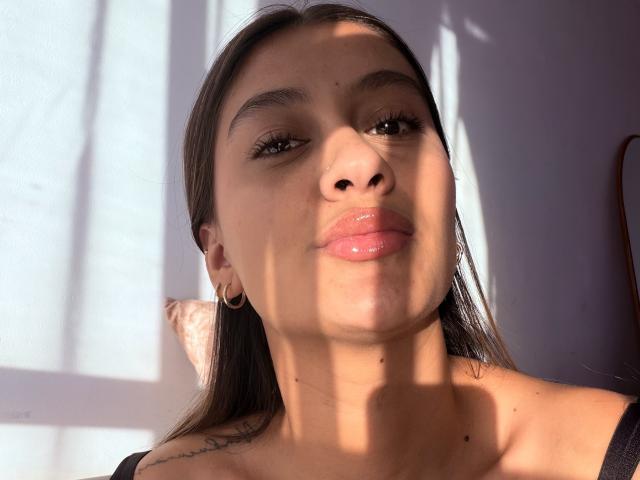 ThaliaAres has glossy pink lips, brown eyes, and long brown hair with sunlight casting shadows on her face. She's wearing gold earrings and a black top.