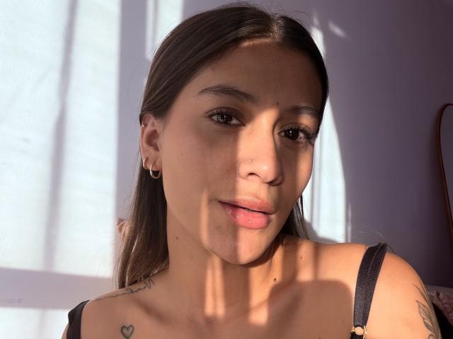 ThaliaAres has light brown skin, long brown hair, and small hoop earrings. Sunlight casts shadows on her face, showing tattoos on her collarbone and upper chest. Boobs visible.