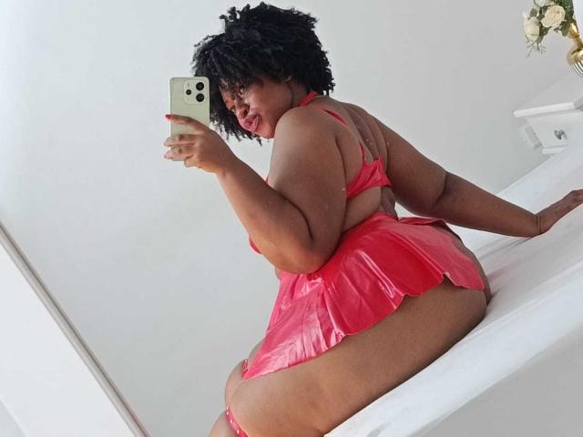 ViaraMoon with dark curly hair, red shiny lingerie, and plump ass, taking a mirror selfie, showing her back and butt, holding a gold phone, sitting on a white bed.