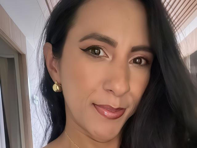 JadeFoster has dark brown eyes, black hair, and glossy pink lips. She's wearing gold earrings and has a subtle smile with winged eyeliner. Her skin is light brown.