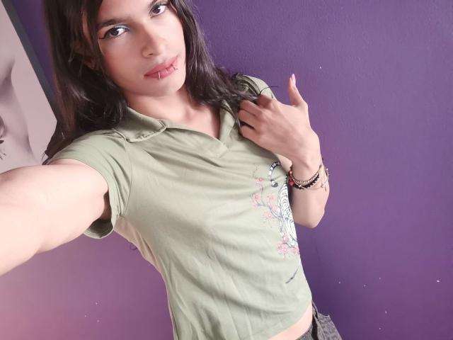LunarisSmith with medium tits, green polo, pink lip ring, dark hair, holding shirt, purple wall, giving a thumbs-up, bracelet on right wrist, selfie, slight smile.