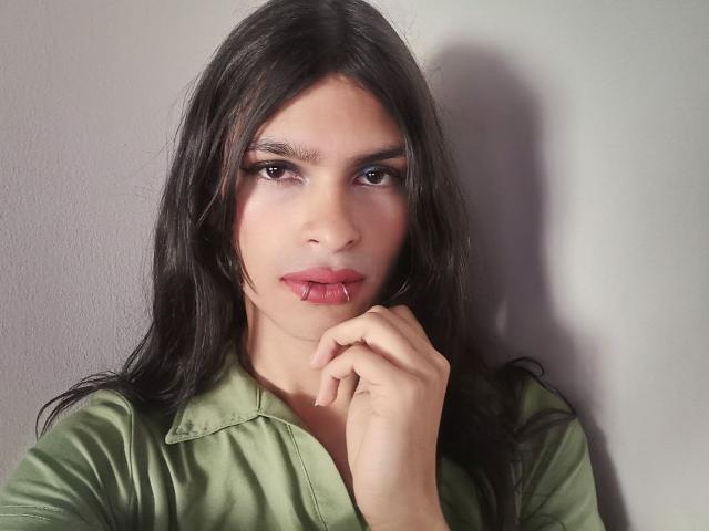 LunarisSmith with long black hair, green shirt, and subtle makeup, touching her slightly parted lips with her finger. Her brown eyes look forward with a soft, pouty expression.
