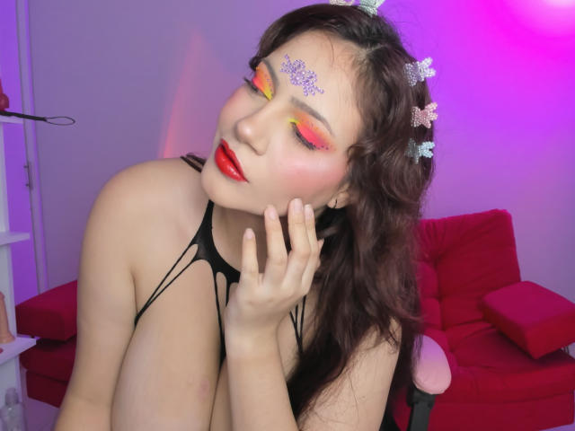 JesicaBomShell with bright red lipstick, colorful eye makeup, and butterfly headpiece, touching her cheek, wearing a black strappy top, exposing her large tits. Purple and pink lights.